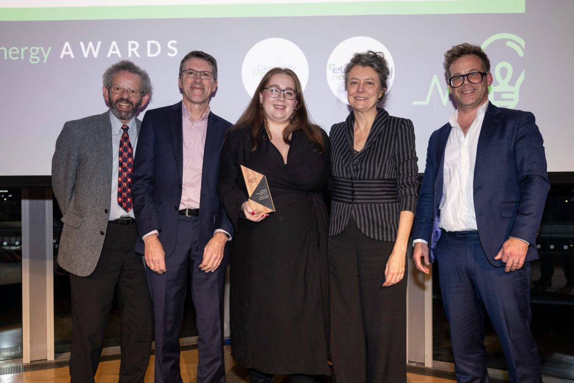 Five people in smart dress stand on a stage at an awards event, with one holding a trophy.