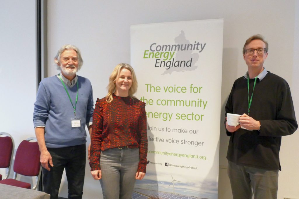 A white haired man, a woman in a stylish autumnal blouse and a bespectacled man holding a cup of tea pose around a Community Energy England pull-up banner.