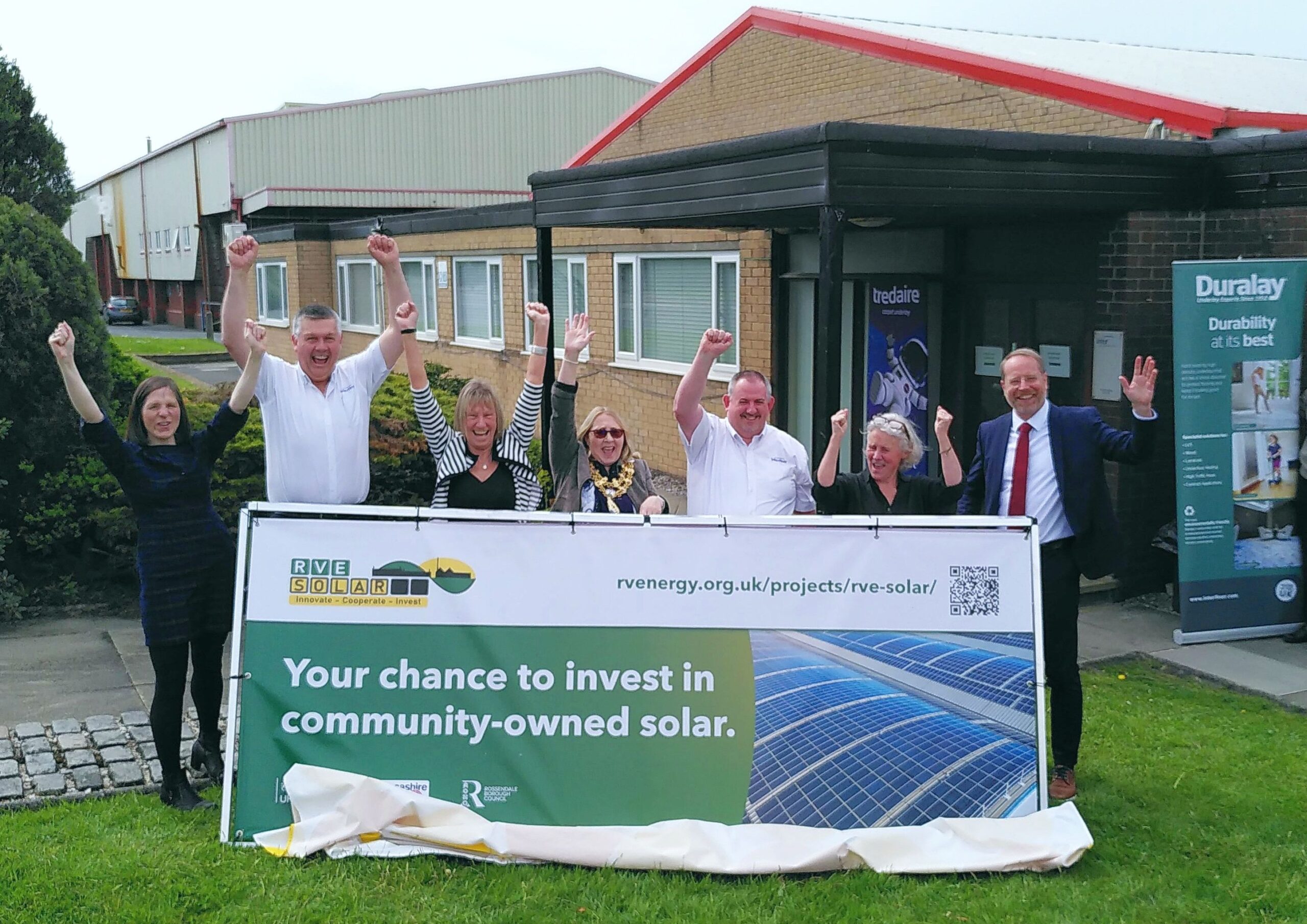 A group of people with arms raised in a cheer stand behind a banner on grass in front of a factory in sunshine. The banner reads "Your chance to invest in community solar".