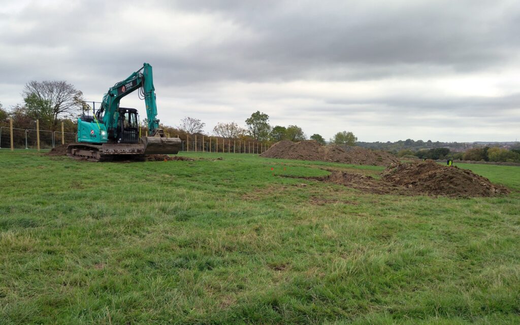 A blue digger at work in a green field which has piles of earth and small flags visible.