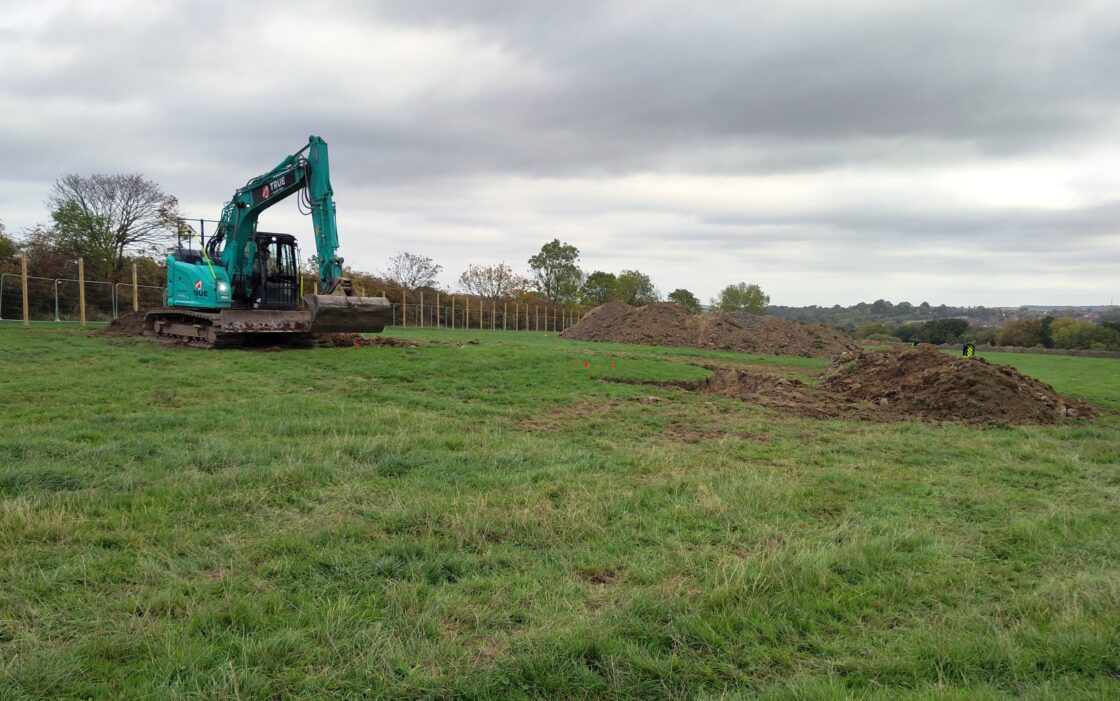 A blue digger at work in a green field which has piles of earth and small flags visible.