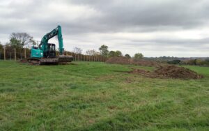 A blue digger at work in a green field which has piles of earth and small flags visible.